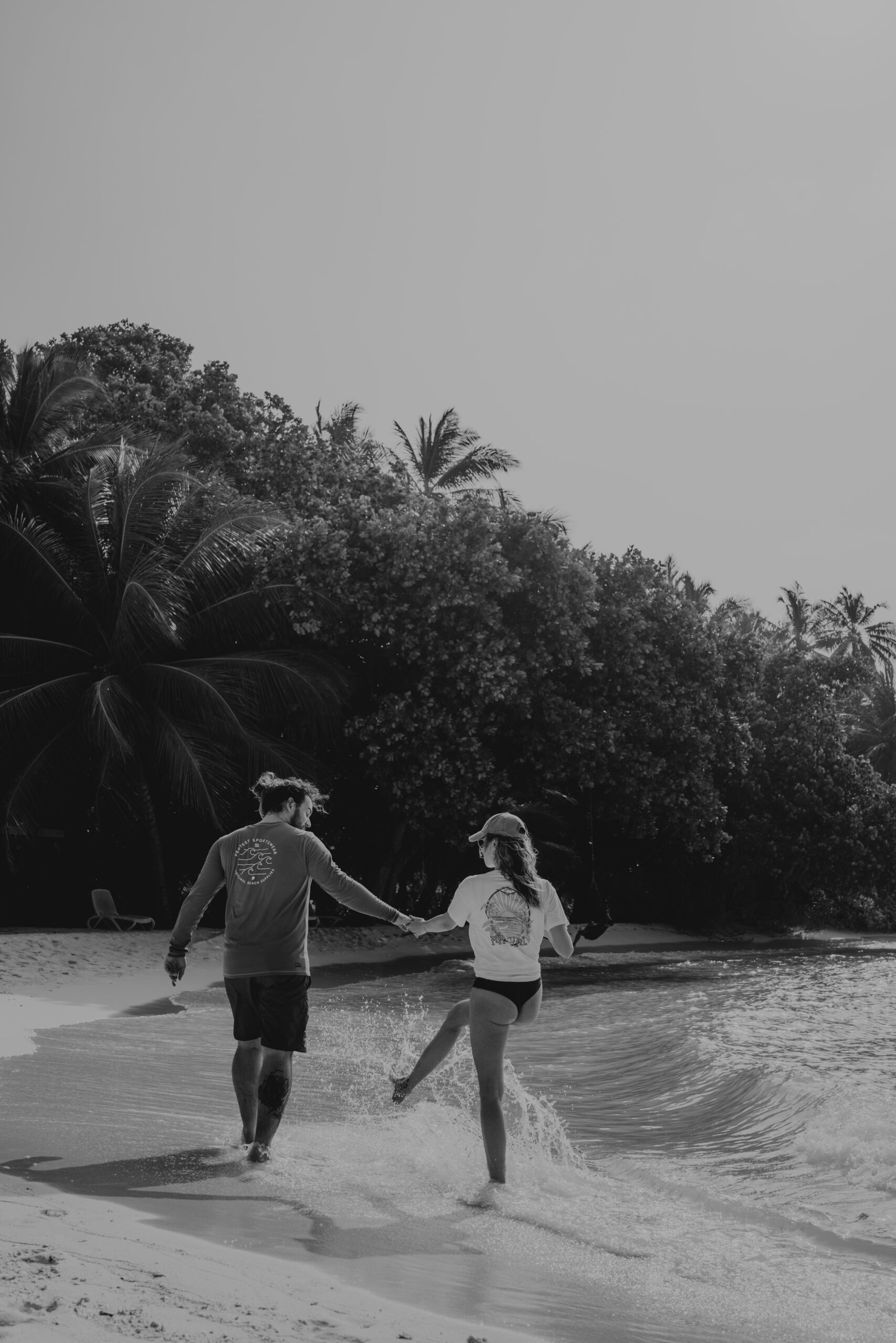 Alex & Katy of Kaiser Studios enjoying a playful beach moment – splashing water, palm trees, and fresh coconuts in a tropical paradise
