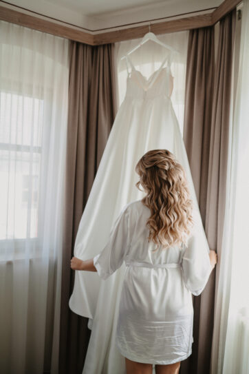 Bride admires her wedding gown at the window – a timeless getting ready moment in Birkenheide