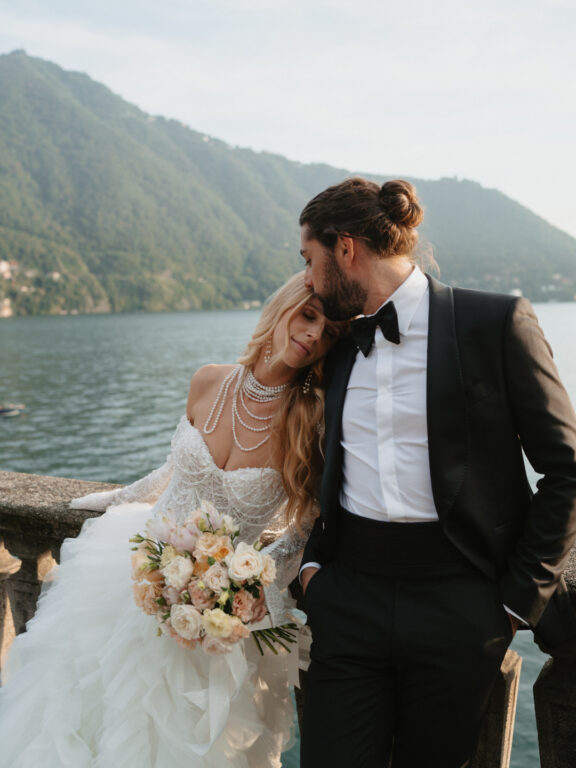 Romantic moment at Villa Pizzo, Lake Como – a luxury wedding couple with a breathtaking view as the groom kisses her forehead.